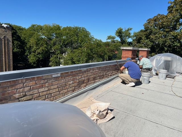 Worker kneeling on a rooftop, inspecting brick parapet and roof membrane under a blue sky.
