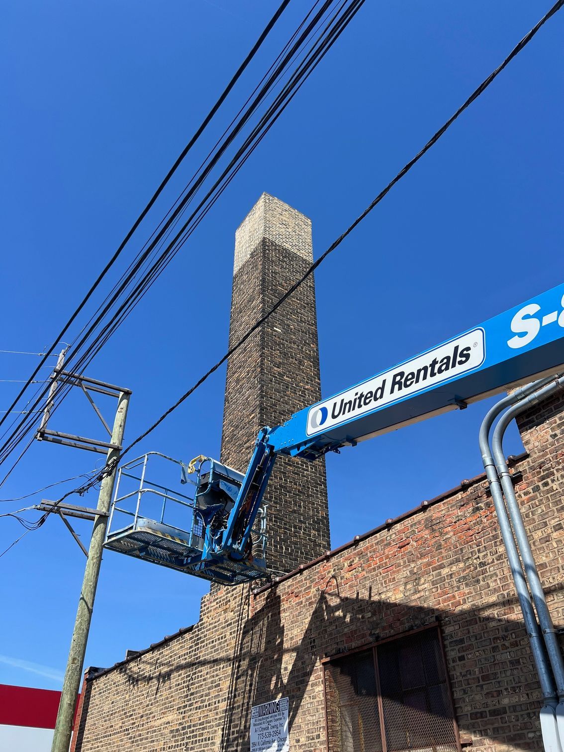 Blue utility lift beside a brick building, reaching toward overhead power lines under a clear blue sky