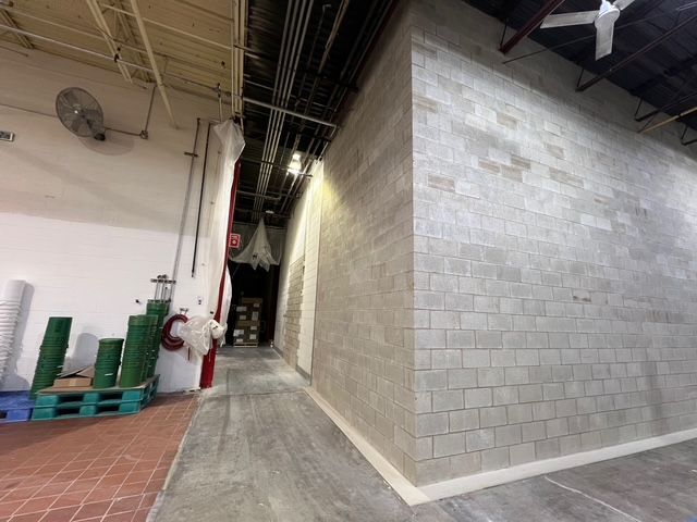 Narrow industrial corridor with gray cinderblock wall, terracotta floor, stacked green chairs, and ceiling pipes