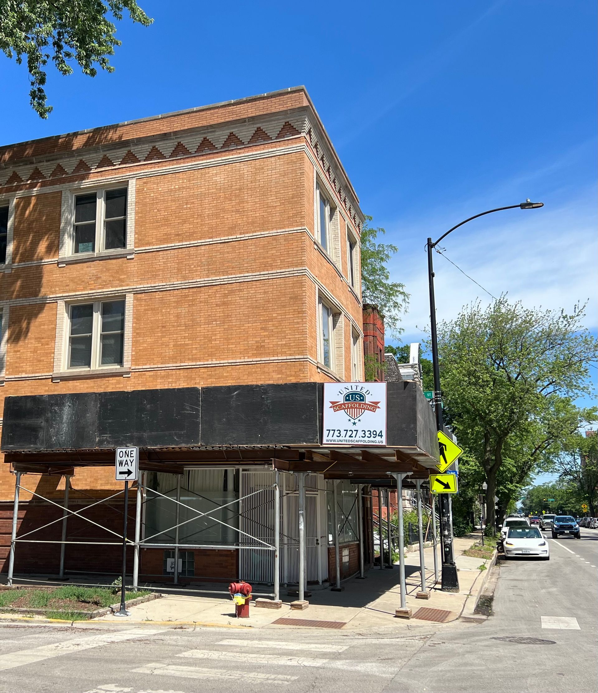 Orange-brick corner building with scaffolding on a sunny street, seen from the sidewalk