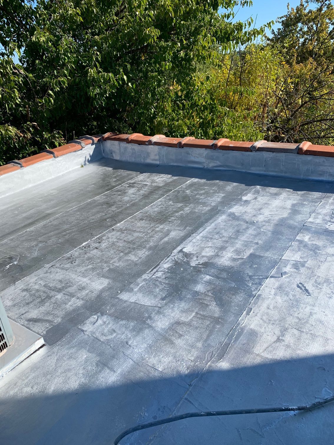 Flat rooftop with gray waterproof coating and red trim, bordered by trees in sunlight