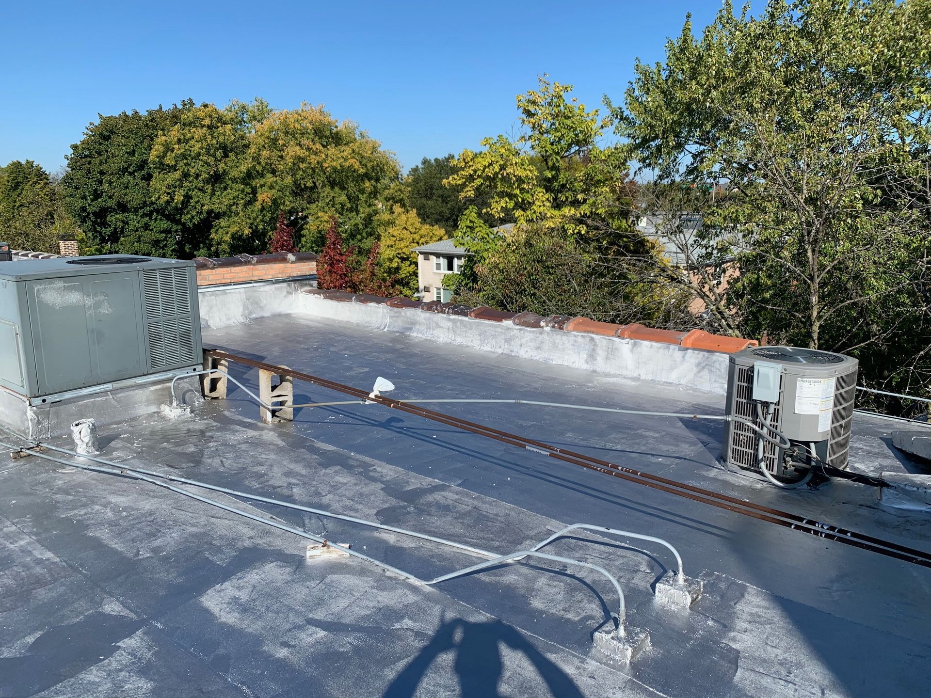 Flat rooftop with HVAC units, vent pipes, and a view of trees and houses beyond.