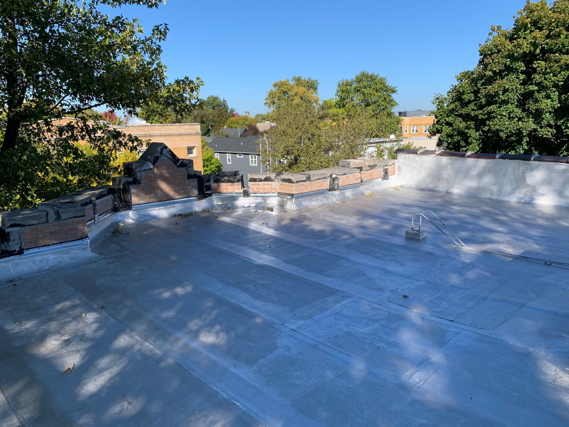 Rooftop under construction with white waterproof coating, brick parapet, and trees against a blue sky