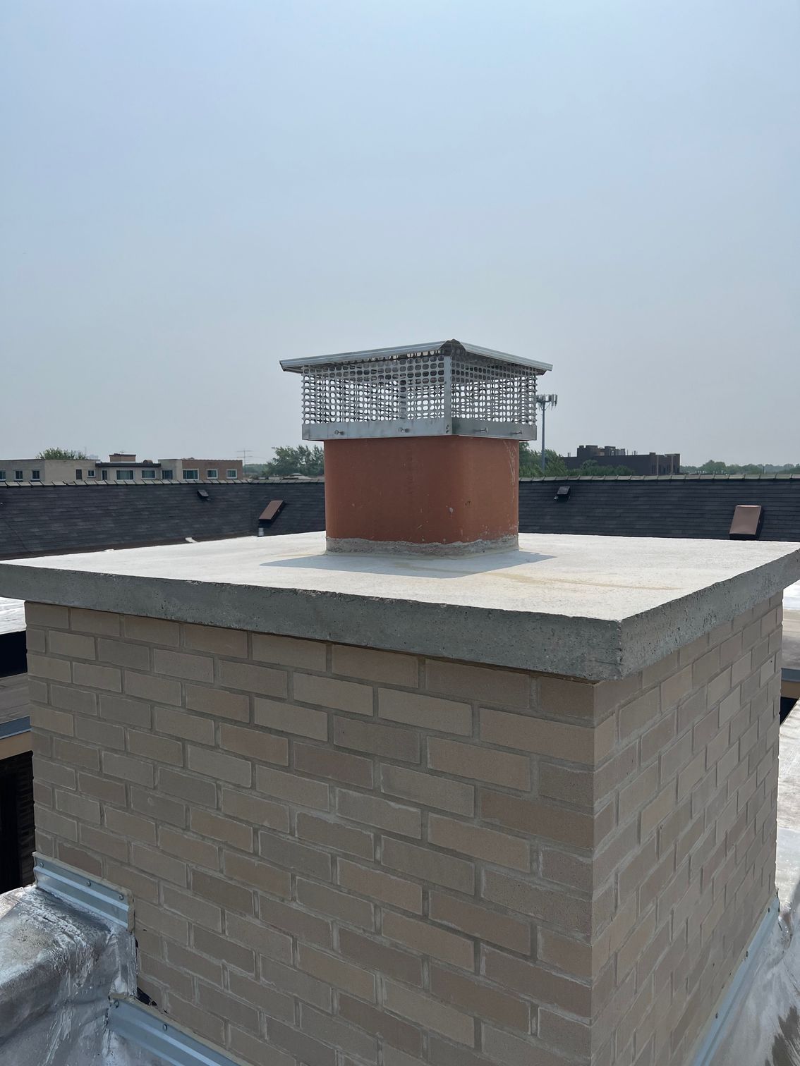 Brick rooftop chimney with a metal vent cap against a hazy sky