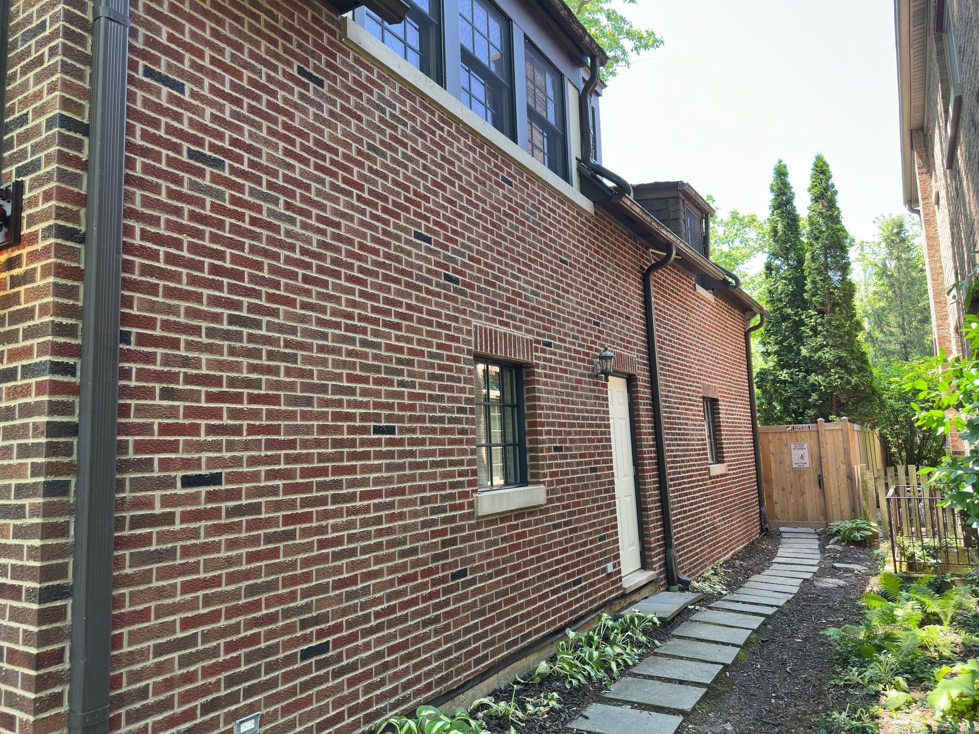 Brick side yard beside a house, with a narrow stone path, windows, and a wooden gate at the end.