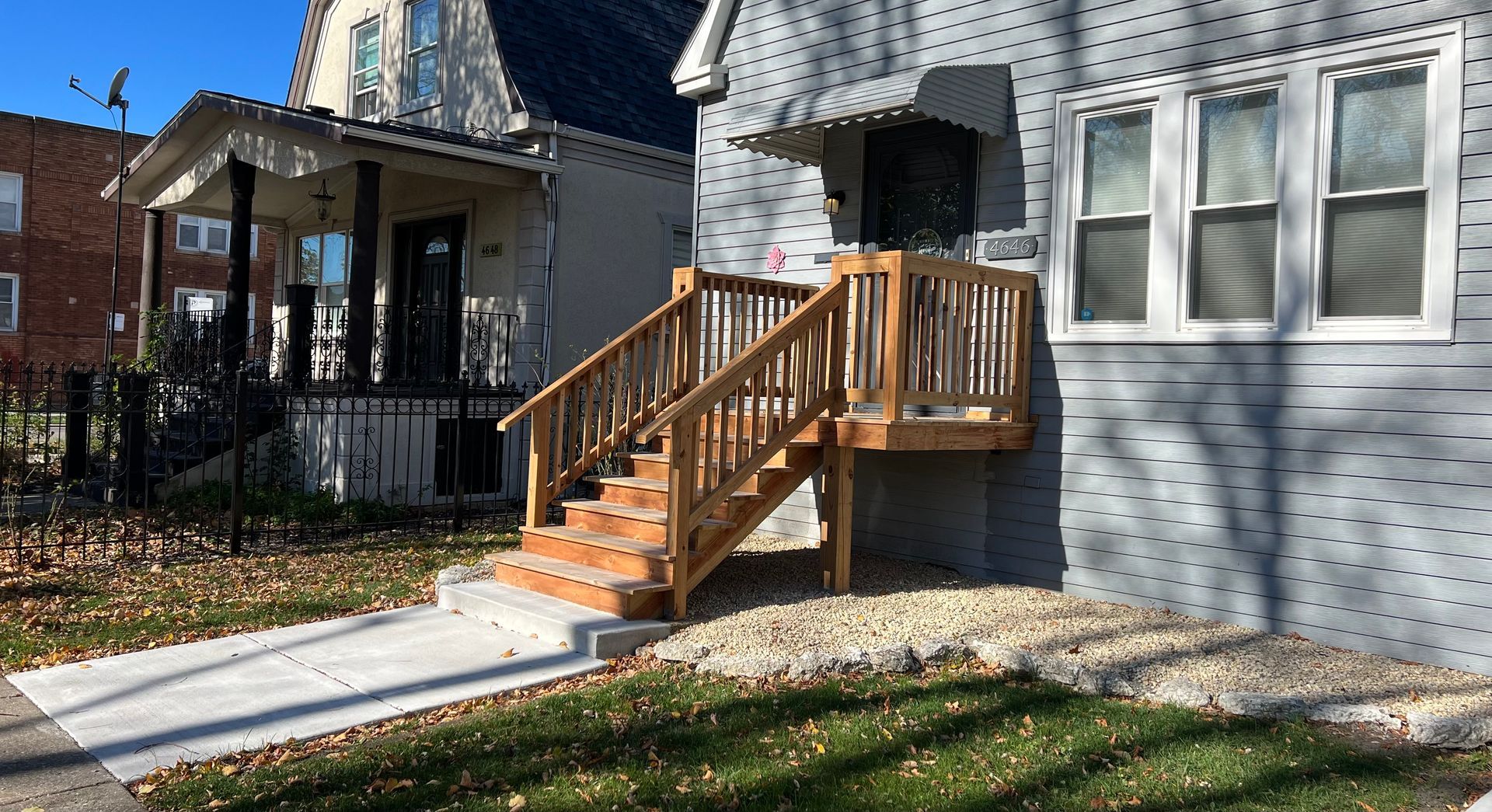 Front porch of a gray house with wooden stairs and railing, beside a sidewalk and yard.