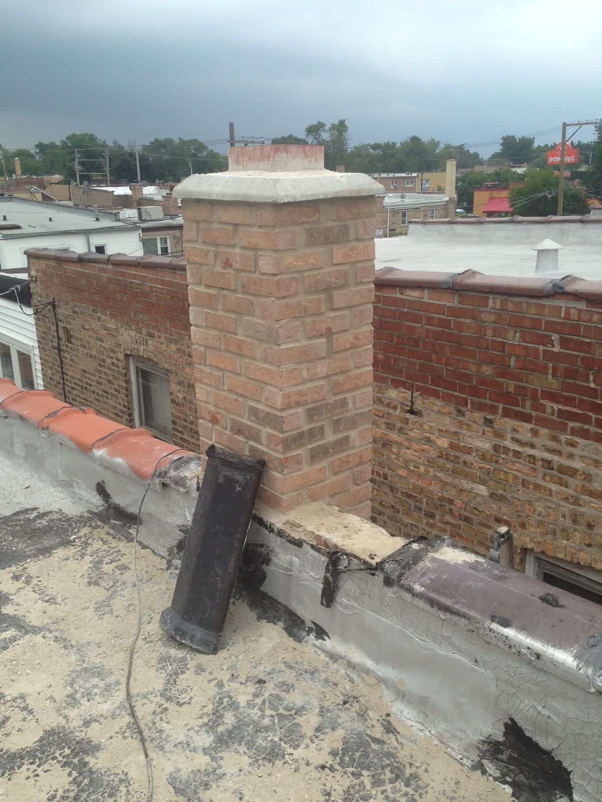Rooftop chimney made of tan brick beside a black vent pipe on a flat roof under overcast sky