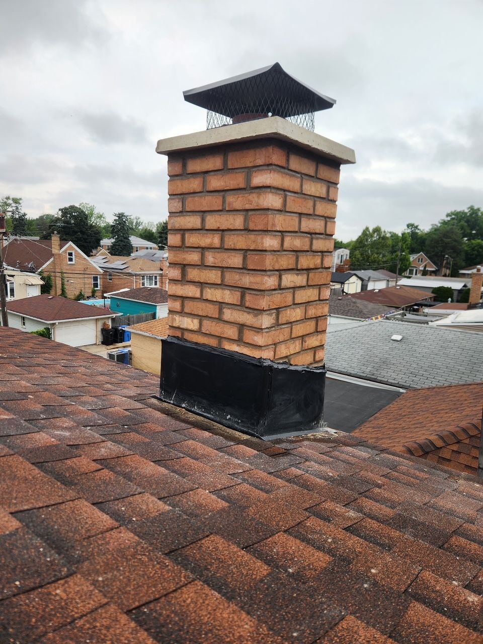 Brick chimney with black cap on a shingled rooftop under a cloudy sky.