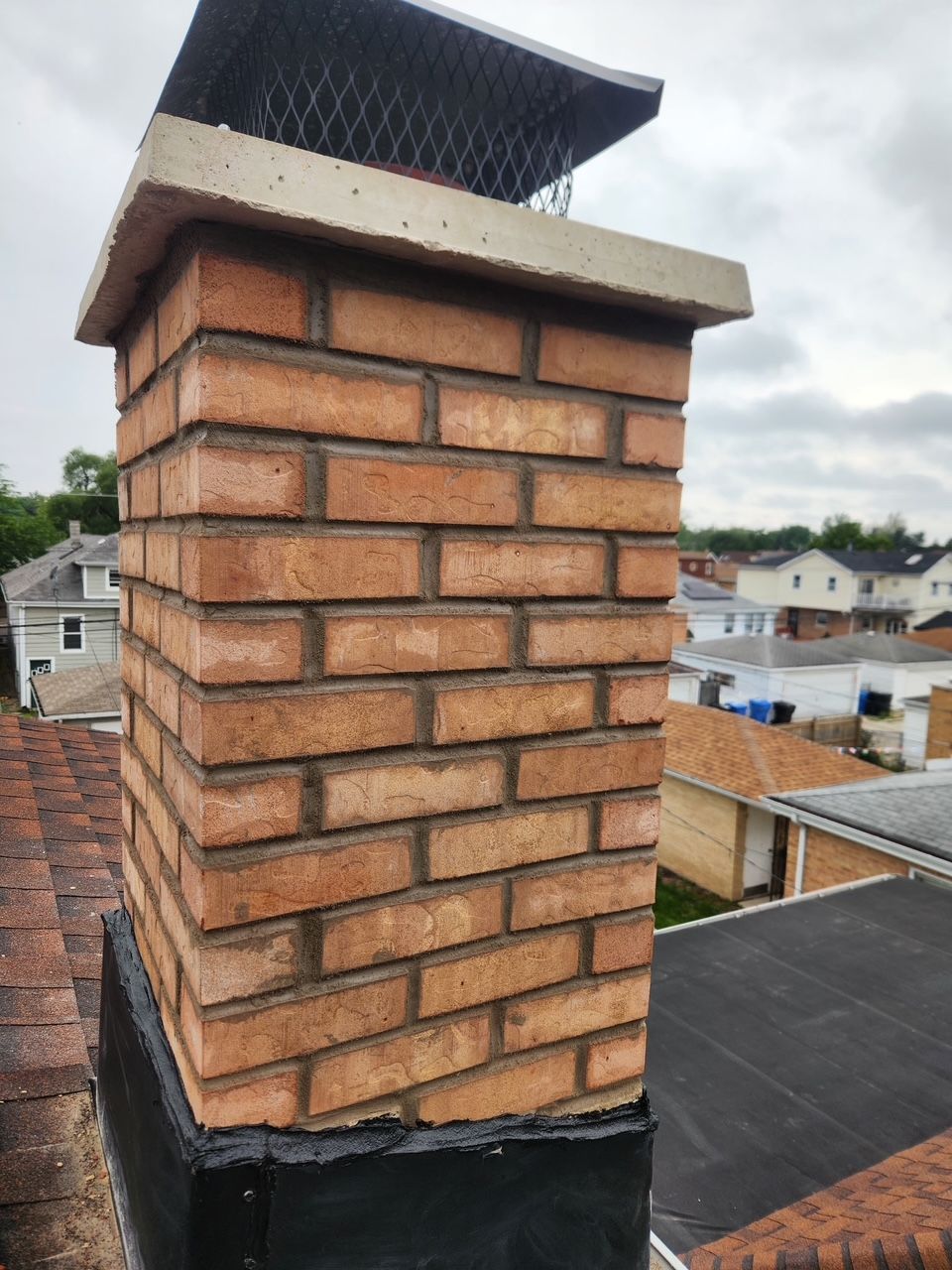 Brick chimney with a metal cap on a rooftop, with houses visible in the background.