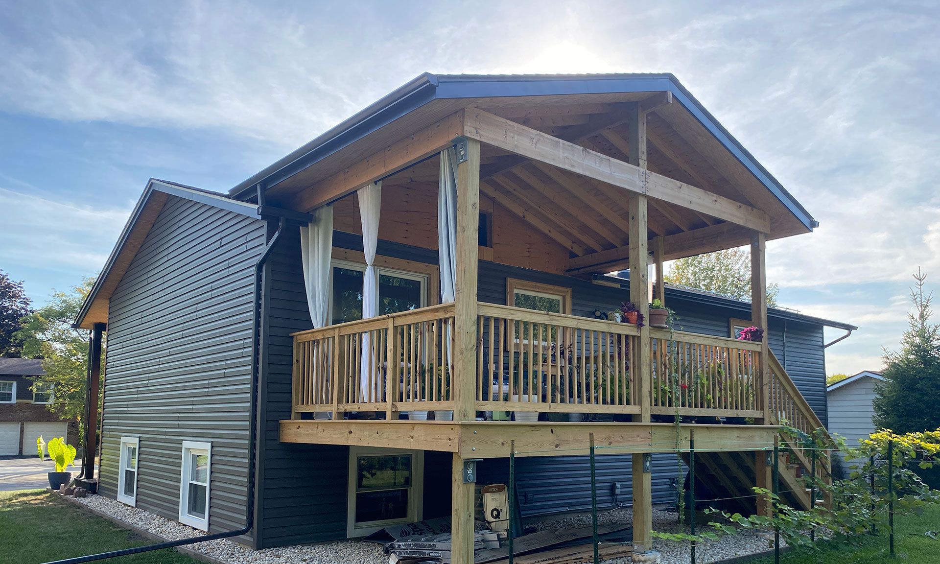 Covered wooden porch with railing on a gray house, viewed from the side in a sunny yard