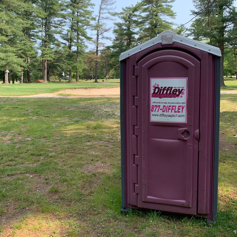 A purple portable toilet is sitting in the grass in a park.