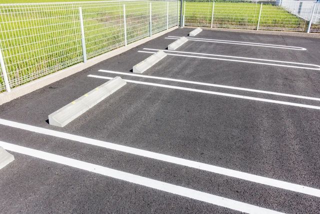 Empty outdoor parking spaces with white lines and concrete wheel stops beside a metal fence.