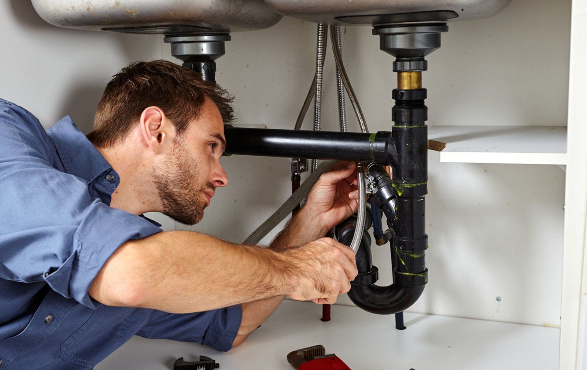 Plumber in blue shirt under a sink, working on pipes with tools.