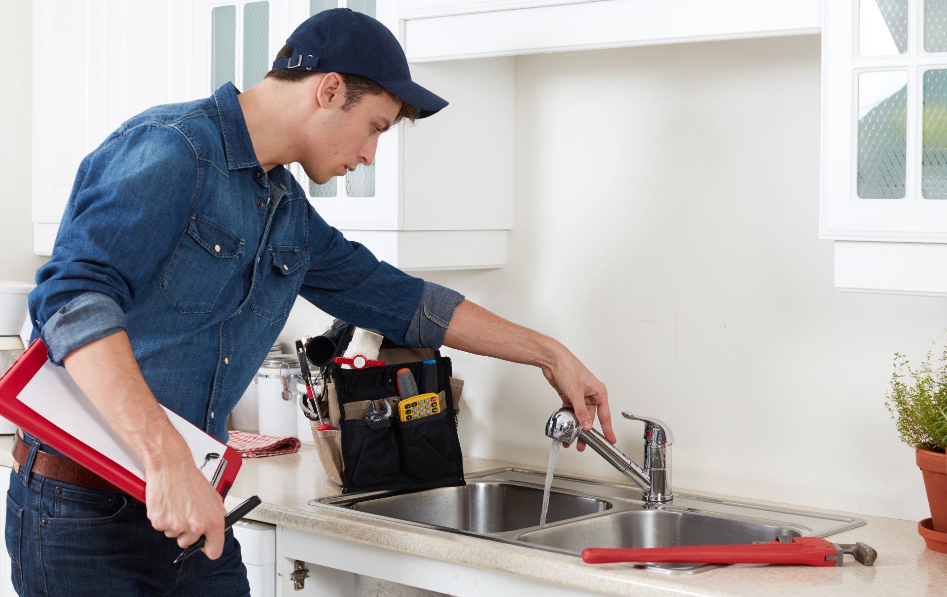 Plumber in blue denim shirt and cap testing a faucet in a kitchen.