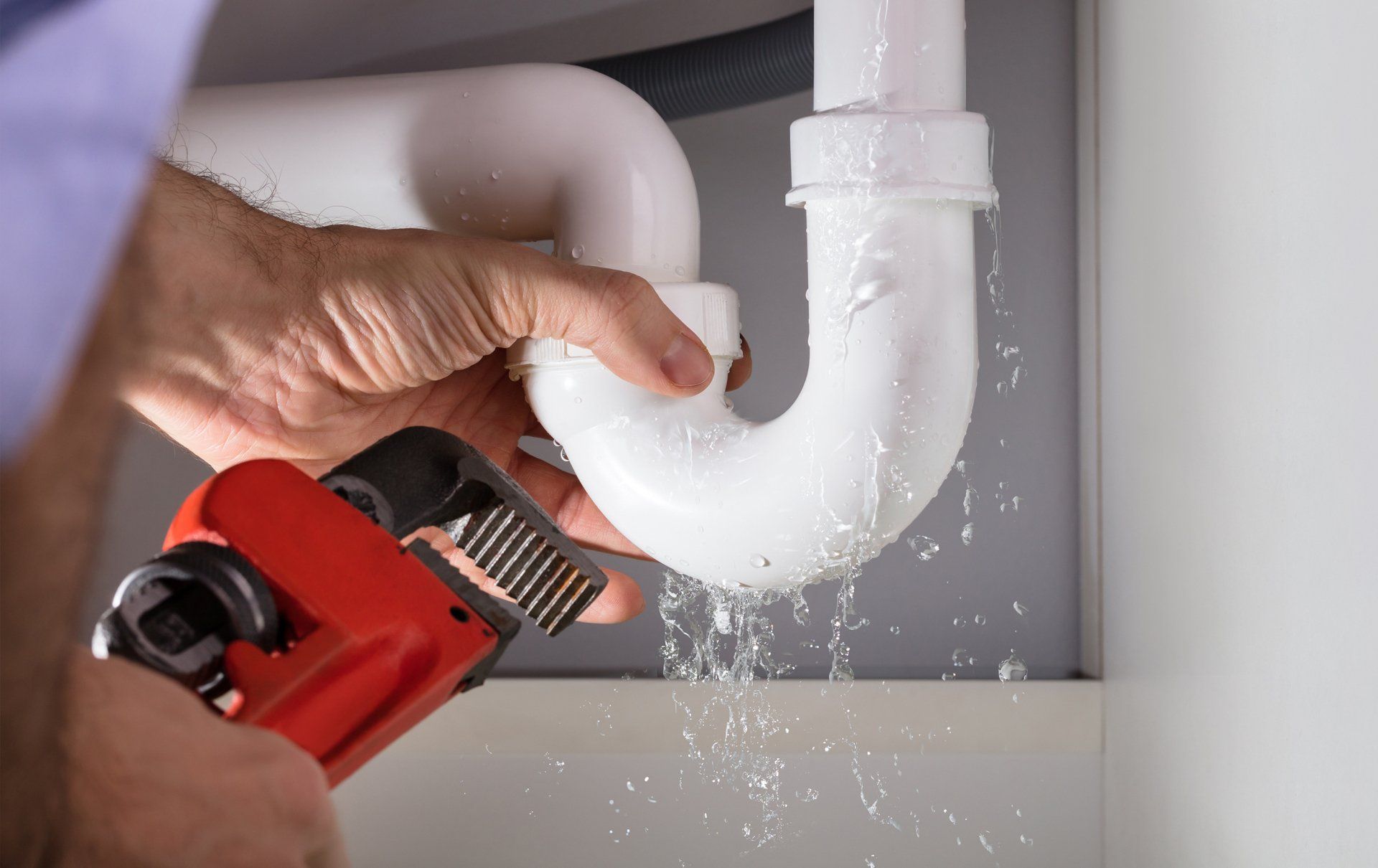 A plumber fixing a leaky white pipe under a sink using a red wrench. Water is spraying.
