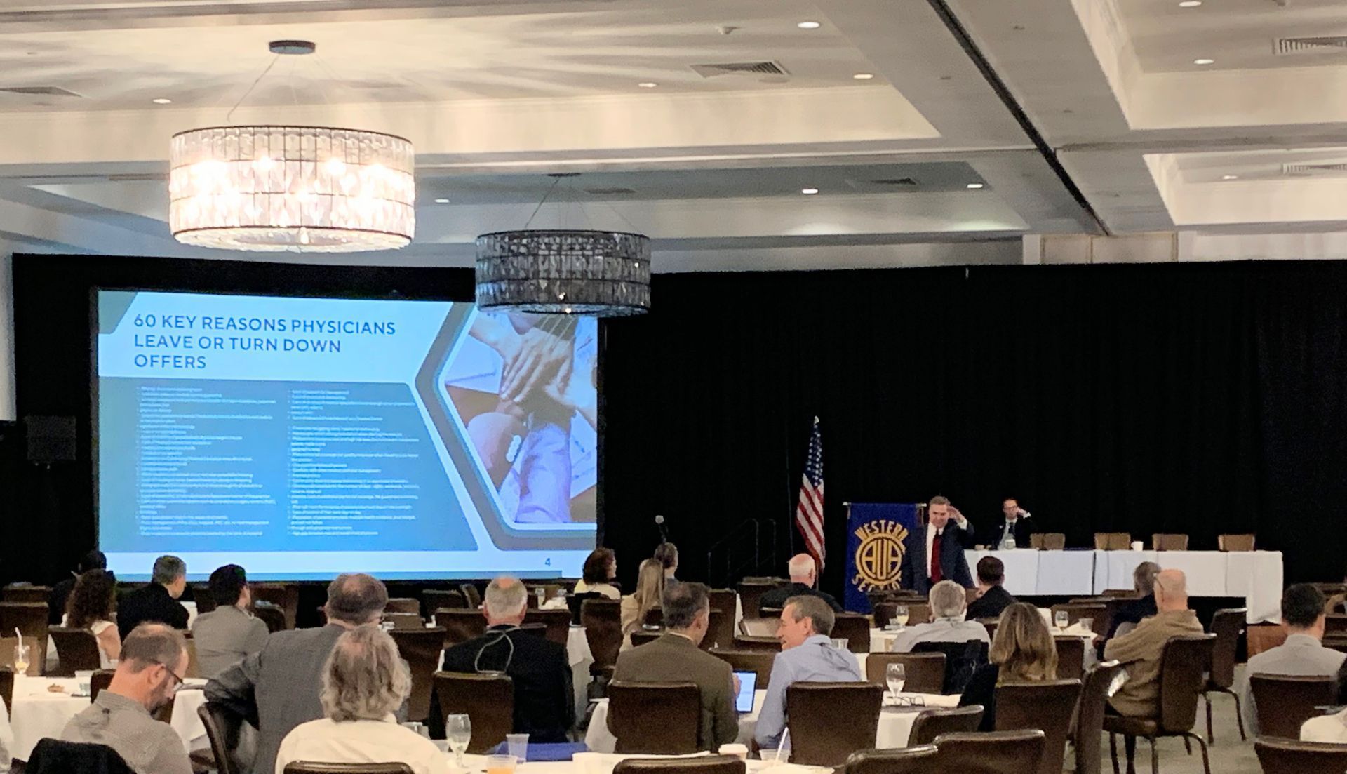 A presentation with a speaker, screen, and audience in a conference setting. Attendees seated at tables.