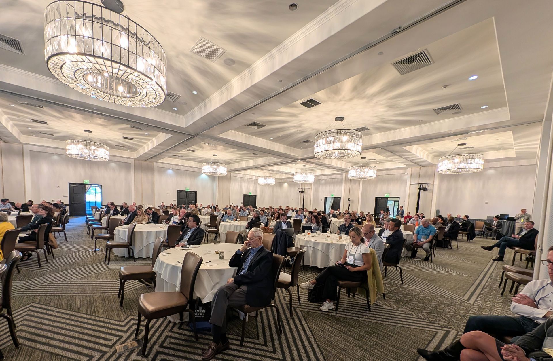 Large room with seated attendees at round tables, under chandeliers.