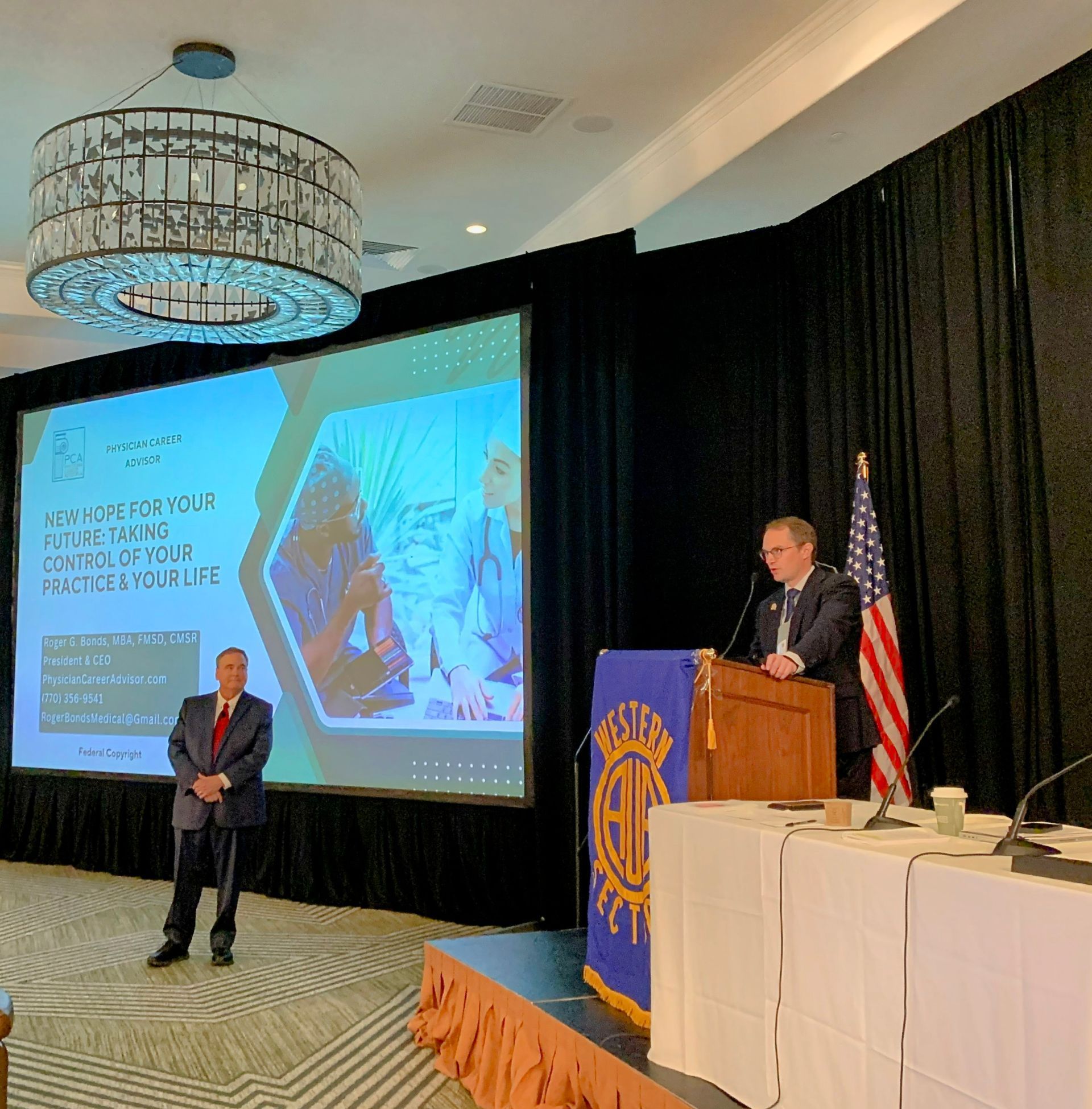 Man speaks at podium, another stands beside a screen with text, American flag present. Presentation setting.