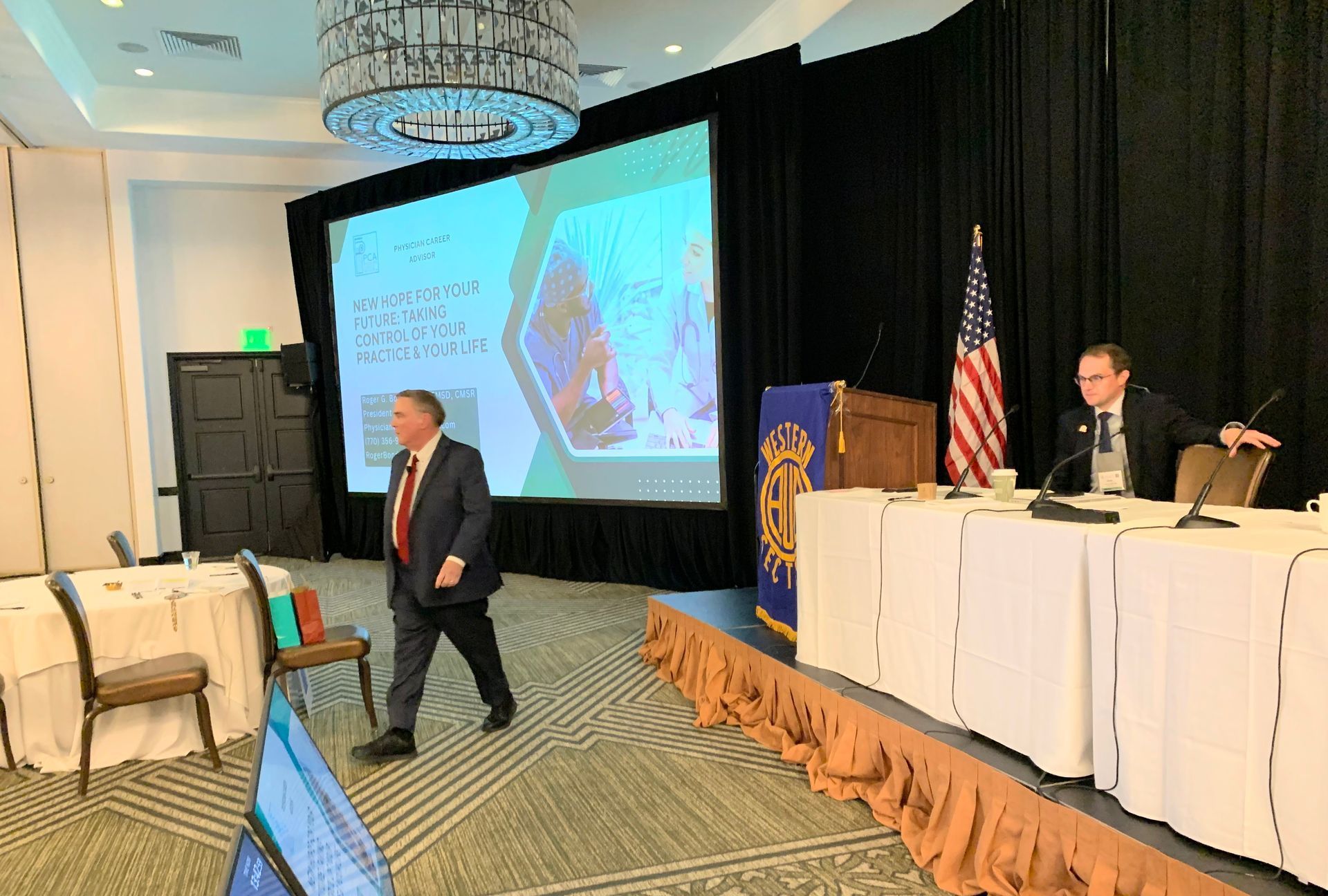 Man walking on stage at conference, another seated at table with microphones, screen showing presentation.
