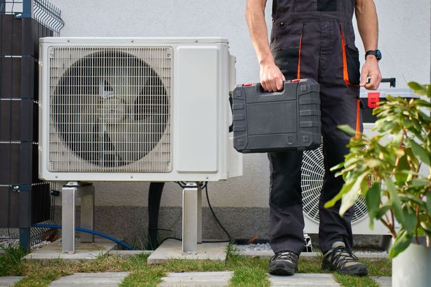 Technician carrying a toolbox beside an outdoor air conditioning unit in a yard.