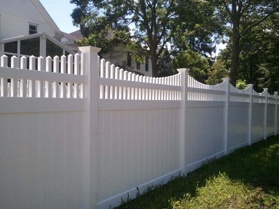 A white vinyl fence with a scalloped picket top design, viewed from the side on a sunny day with trees in the background.