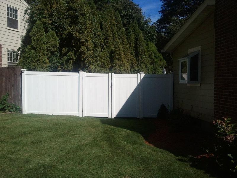 A white vinyl privacy fence with a double gate in a backyard between a house and a large tree line.