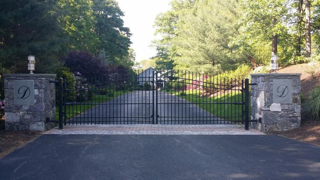 A closed black metal driveway gate stands between two stone pillars featuring the letter 