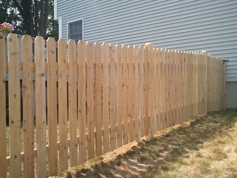 A new, light-colored wooden picket fence runs along the side of a grey-sided house in a grassy yard.
