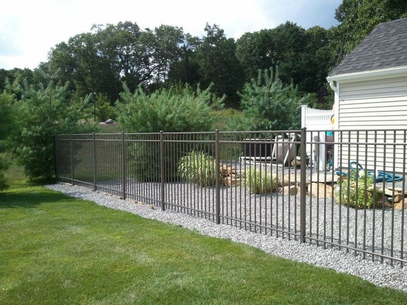 A metal fence lines a gravel bed beside a house, bordering a green lawn with trees and shrubs in the background.