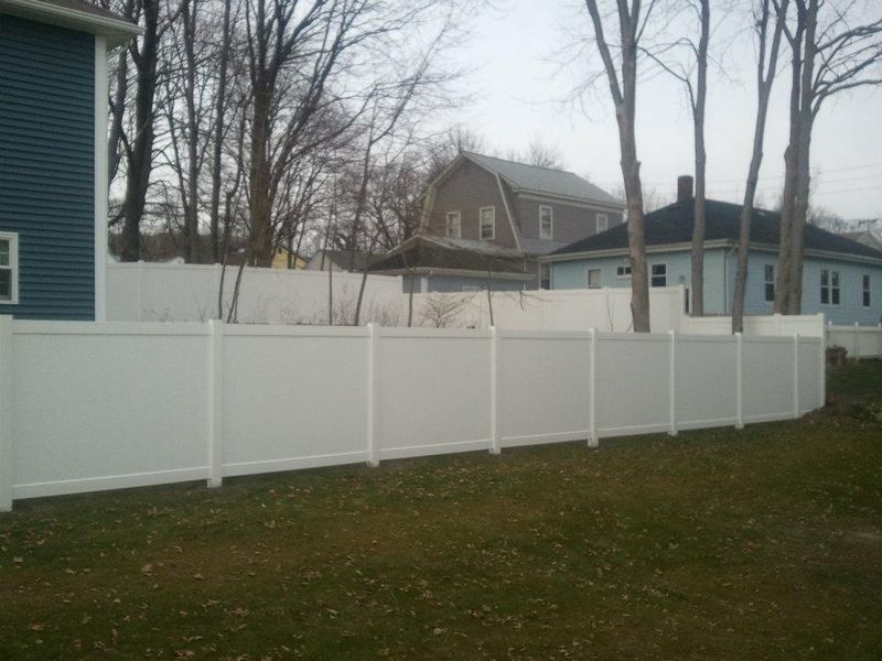 A tall, white vinyl privacy fence encloses a grassy backyard, with houses and trees visible in the background.