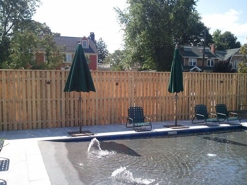 A wooden privacy fence surrounds a pool with two closed green umbrellas and lounge chairs on a sunny patio.