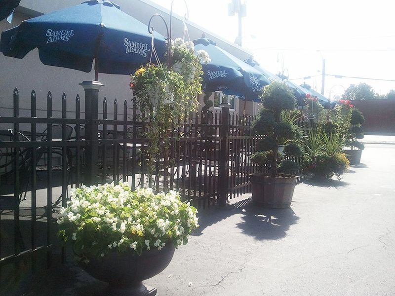 Outdoor patio seating with dark metal fencing, blue umbrellas, and potted flowers lining a sidewalk.