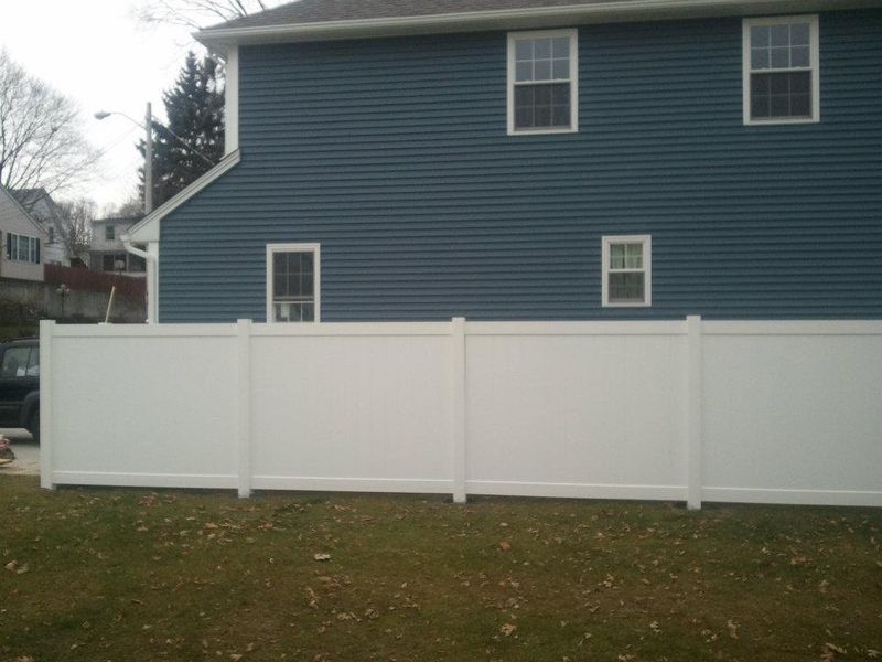 A tall white vinyl privacy fence stands in front of a house with blue siding and white-framed windows on a grassy lawn.