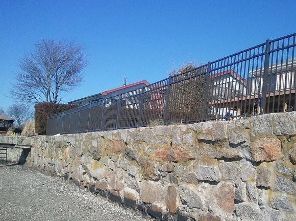 A stone retaining wall topped with a dark metal fence sits along a gravel path beneath a clear blue sky.