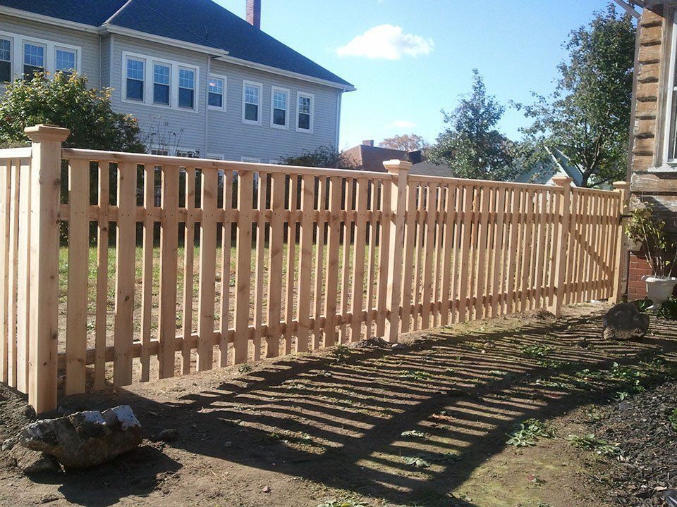 A newly installed light-colored wood picket fence runs across a residential backyard on a sunny day.