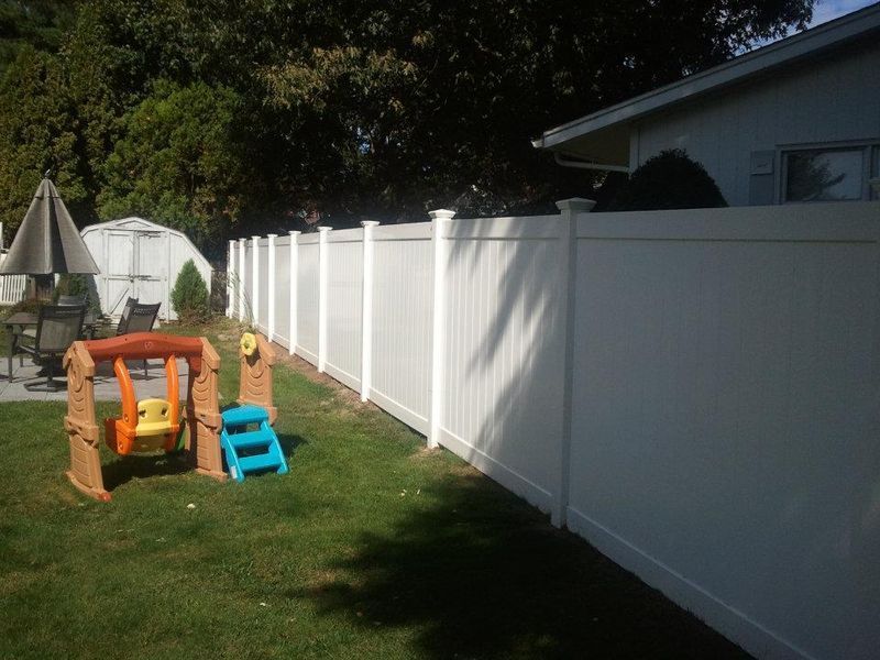 A backyard scene featuring a white vinyl privacy fence, a small children’s swing set, and a patio umbrella on a lawn.