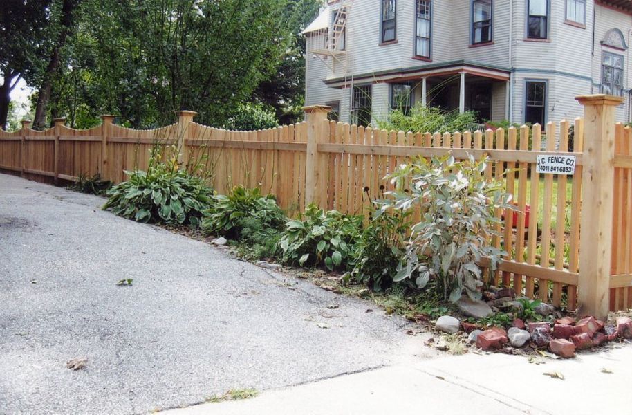 A wooden picket fence with a scalloped top edge runs along a paved driveway next to a house and garden beds.
