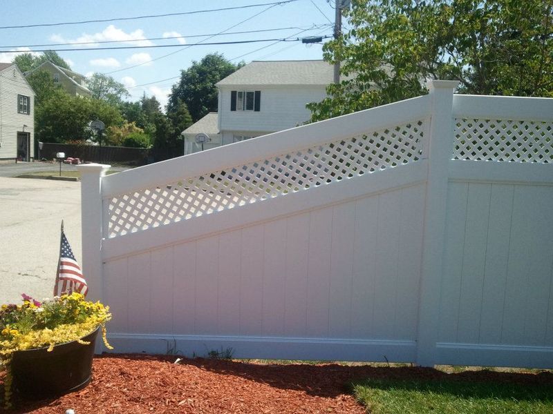 A white vinyl privacy fence with a diagonal lattice topper stands near a house, with a small flag and flowers in the front.