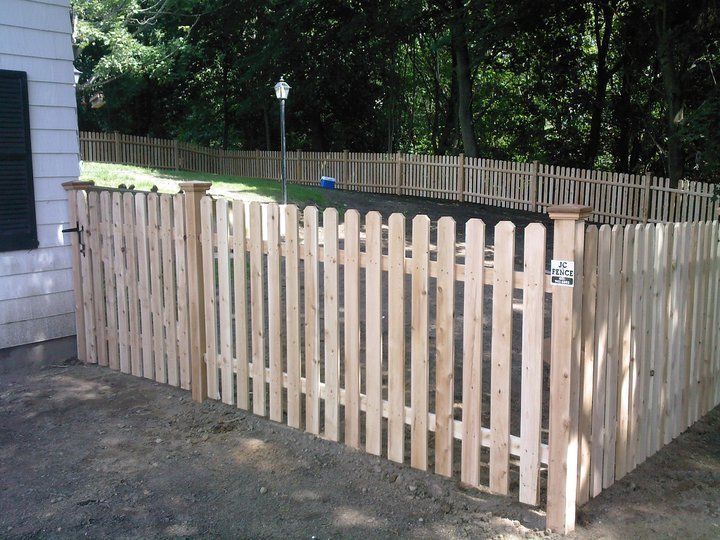 A new, light-colored wooden picket fence borders a residential driveway with a grassy area and trees in the background.