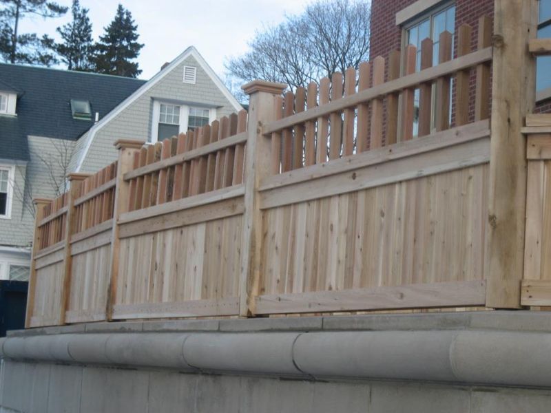 A wooden fence with a picket top, installed atop a light-colored masonry wall in front of a house.