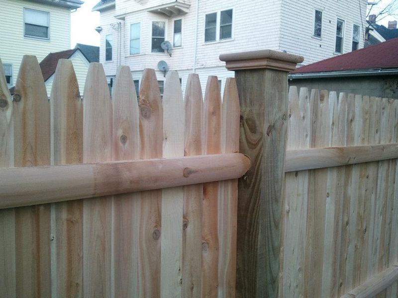 A wooden picket fence featuring a central post with a decorative cap, set against a blurred background of houses.