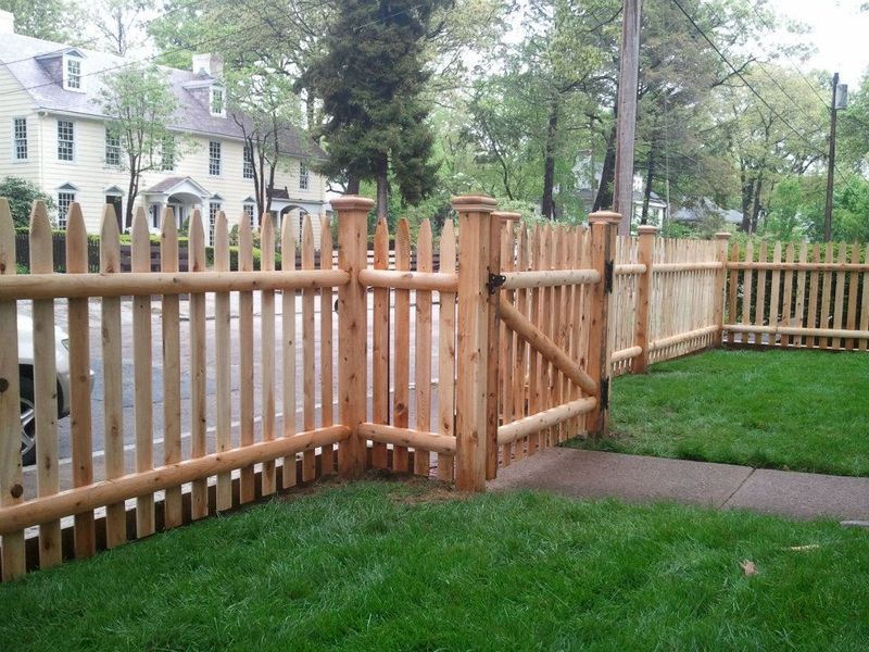 A wooden picket fence with a gate, installed along a green lawn in front of a house on a sunny day.
