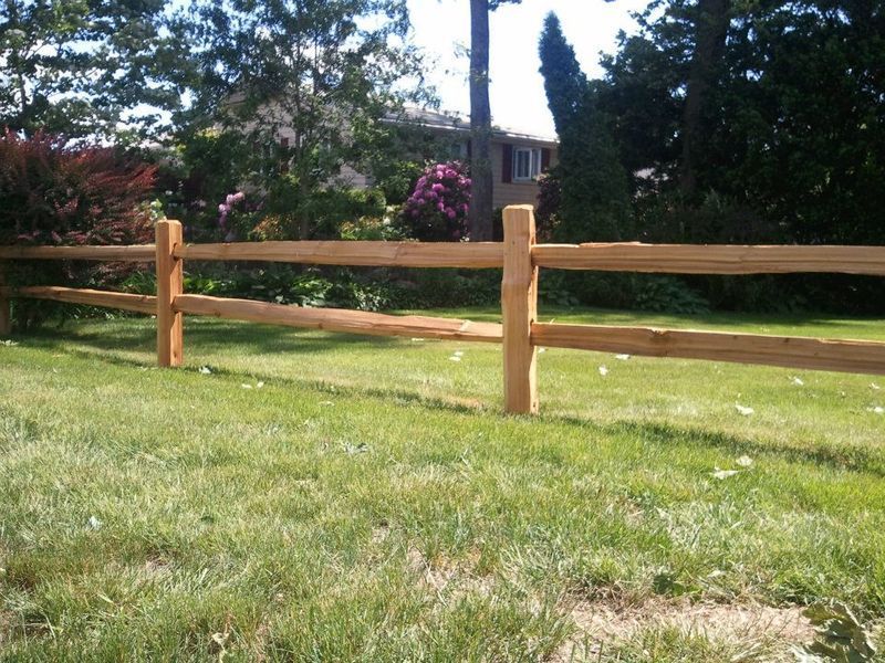 A two-rail split rail wooden fence runs across a green, sunlit lawn in front of a residential home with trees.