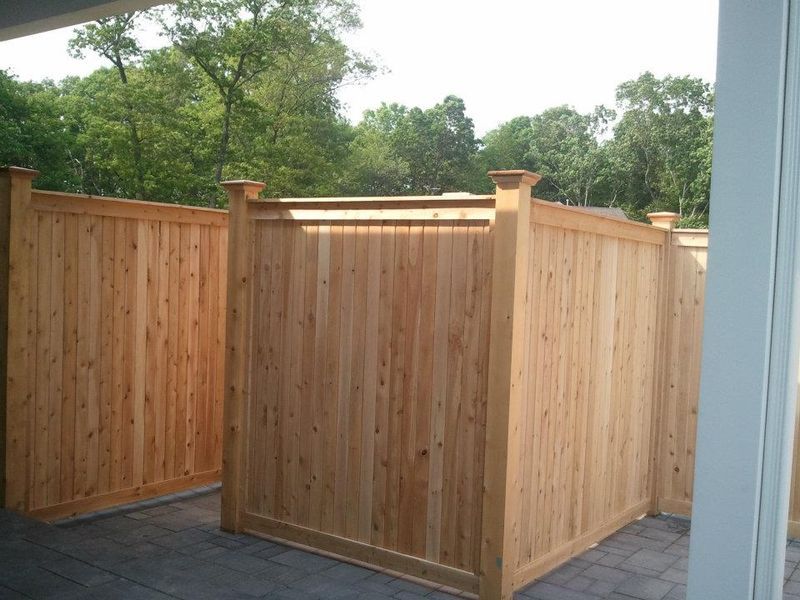 A wooden privacy fence enclosure surrounds a gray paver patio with a leafy green treeline in the background.