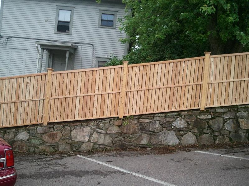 A light wood fence sits atop a stone retaining wall in front of a gray house and a parking space.