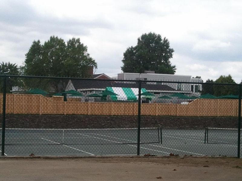 A chain-link fence in front of tennis courts, with a wooden fence and a green and white striped tent in the background.