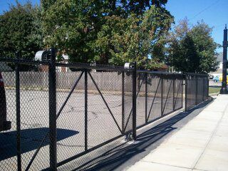 A black chain-link rolling gate along a sidewalk next to a parking lot and trees under a clear blue sky.