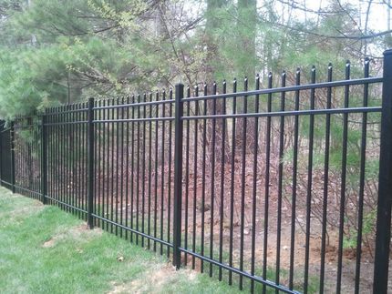 A black, ornamental metal picket fence stands on a grassy lawn in front of a wooded area.