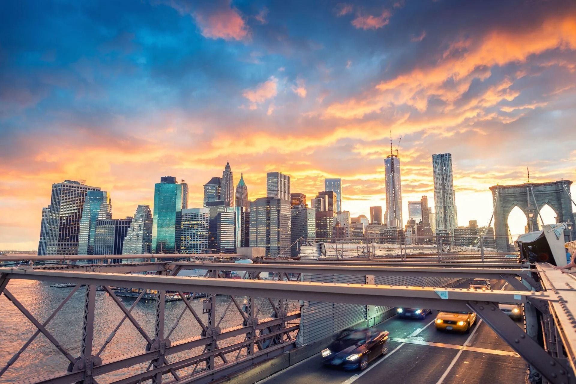 New York City skyline at sunset, viewed from Brooklyn Bridge. Cars drive across bridge, colorful sky.