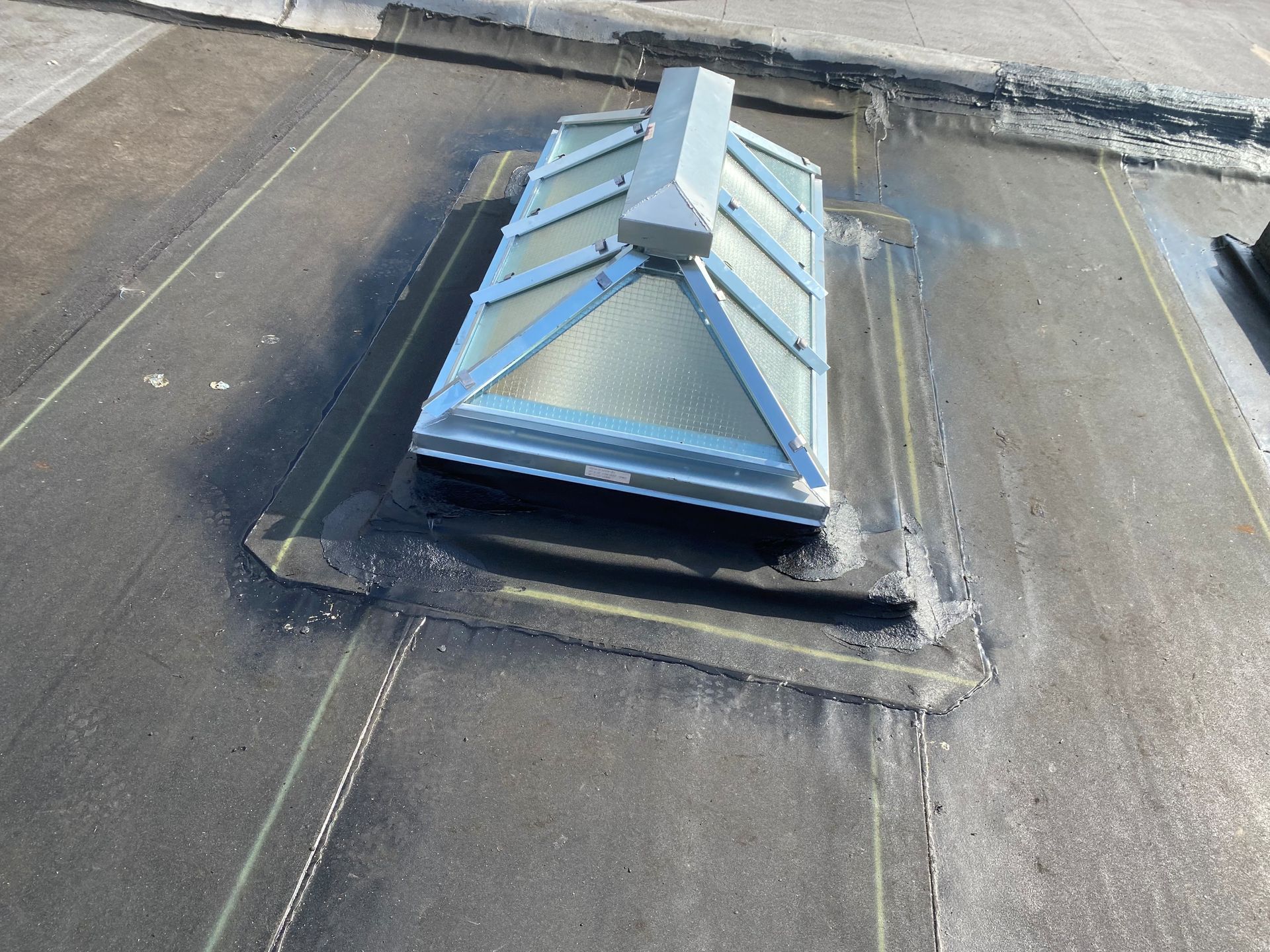 Skylight on a dark roof with black tar sealing around it. The sky light has a light silver frame and multiple glass panels.